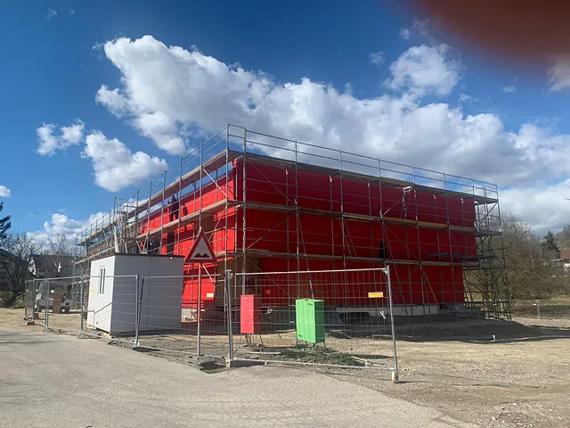 Neubau des Feuerwehr Gästehauses in roter Fassade mit Gerüst und Bauzaun bei sonnigem Himmel