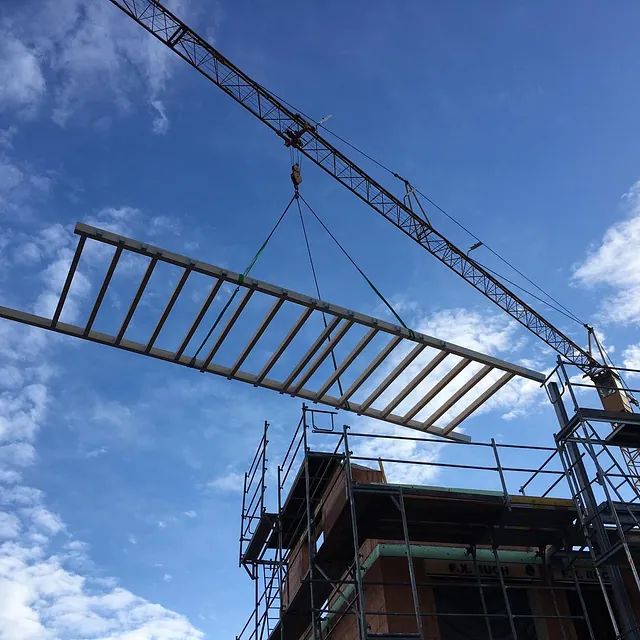 Kran hebt Bauteil über Gerüst eines Mehrfamilienhaus-Neubaus bei blauem Himmel