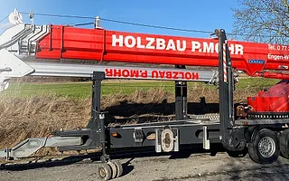 Roter Kran mit weißer Aufschrift 'HOLZBAU P. MOHR' auf dem Ausleger, montiert auf einem schwarzen Fahrzeugrahmen, im Freien vor Büschen und blauem Himmel