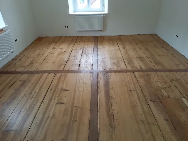 Renovierter Holzboden mit breiten Dielen und sichtbaren Fugen in einem hellen Raum mit weißen Wänden und zwei Heizkörpern unter den Fenstern, Altbausanierung