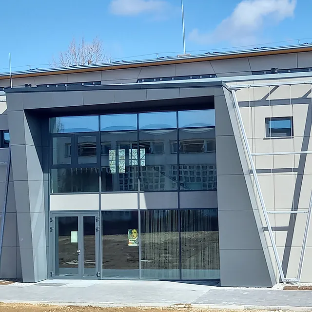Eingangsbereich der Sporthalle in Engen mit moderner Fassade aus grauen Paneelen und großen Glasflächen, Baustelle mit noch unbefestigtem Boden