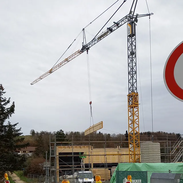 Baukran hebt Holzelement über Baustelle des Feuerwehrgerätehauses in Konstanz-Dettingen, umgeben von Gerüsten und Baustellenfahrzeugen