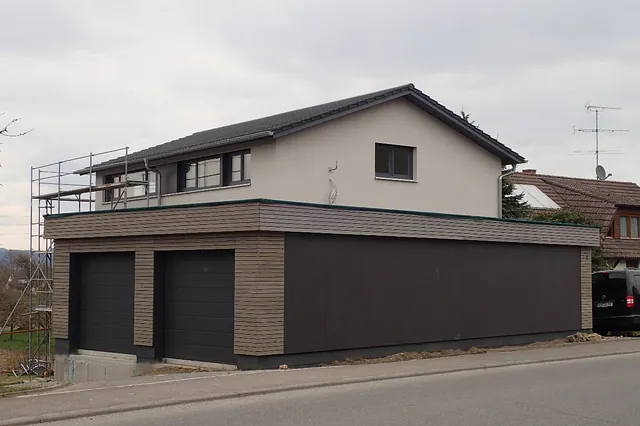 Moderne Hausfassade mit grauem Putz, zwei schwarzen Garagentoren und Holzverkleidung an der unteren Etage, Gerüst an der Seite